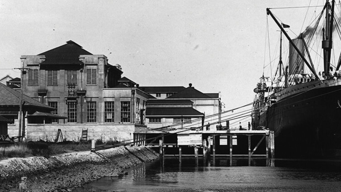 A large ship docked beside a historic building on a calm river, with smokestacks releasing steam. The scene is black and white, giving it a vintage appearance.