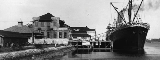 A large ship docked beside a historic building on a calm river, with smokestacks releasing steam. The scene is black and white, giving it a vintage appearance.