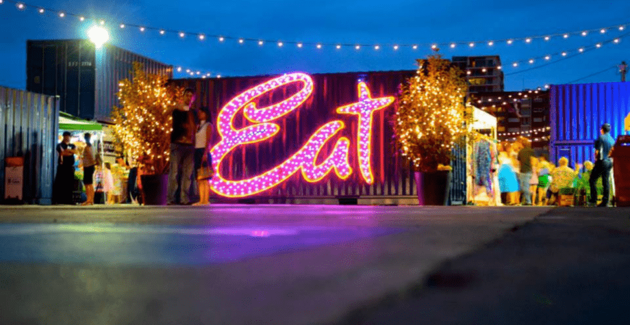 Neon-lit Eat sign at an outdoor night market. String lights hang above, illuminating stalls and people in the background.