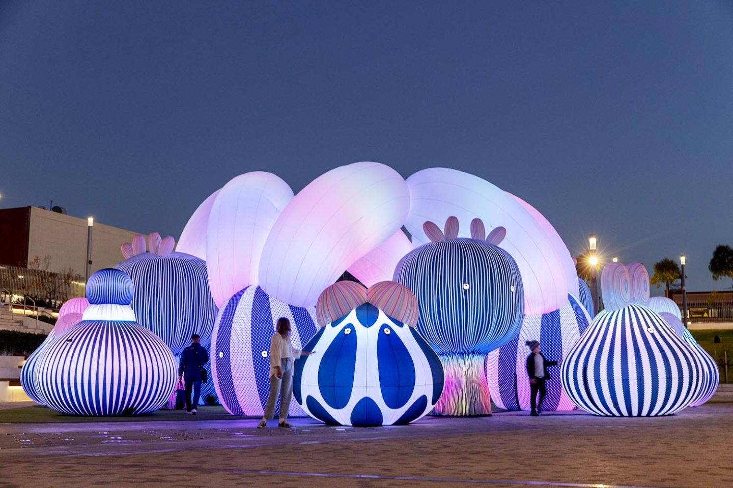 The image shows large, inflatable art installations resembling abstract, multicolored mushrooms with stripes and dots, illuminated in pink, blue, and white hues. People interact with the structures at dusk on a paved area.