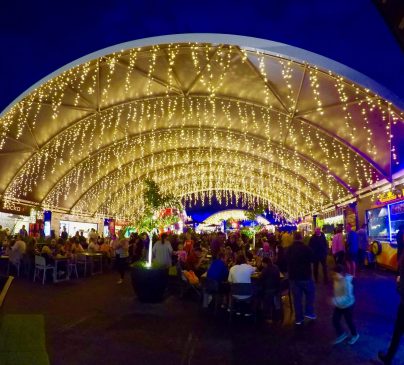 A bustling night market under a large, illuminated canopy adorned with strings of yellow lights. People gather around food stalls and tables, enjoying the lively atmosphere and vibrant surroundings.
