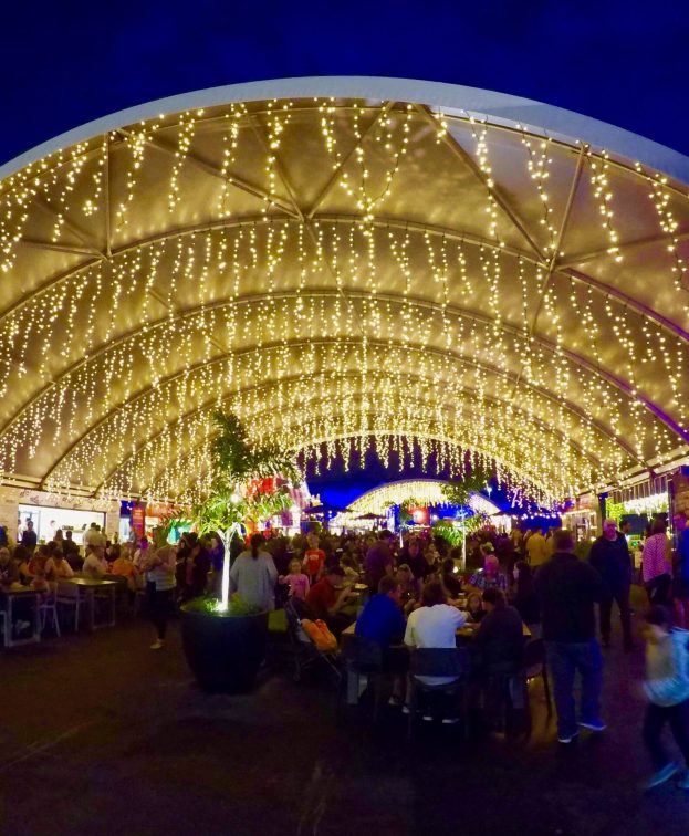 A bustling night market under a large, illuminated canopy adorned with strings of yellow lights. People gather around food stalls and tables, enjoying the lively atmosphere and vibrant surroundings.