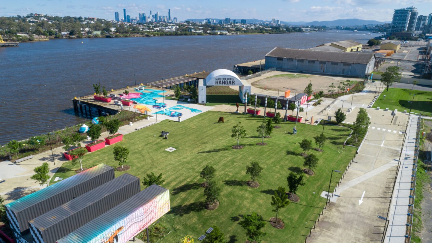 Aerial view of a riverside park with green lawns and scattered trees, showing the Maritime Green Hangar structure.