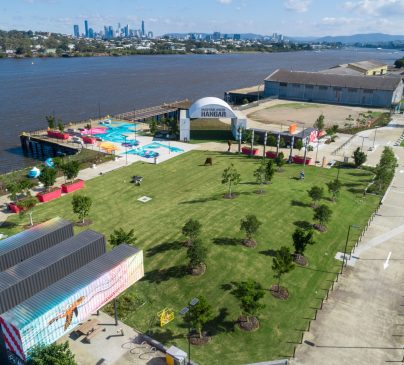 Aerial view of a riverside park with green lawns and scattered trees, showing the Maritime Green Hangar structure.