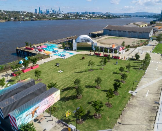 Aerial view of a riverside park with green lawns and scattered trees, showing the Maritime Green Hangar structure.