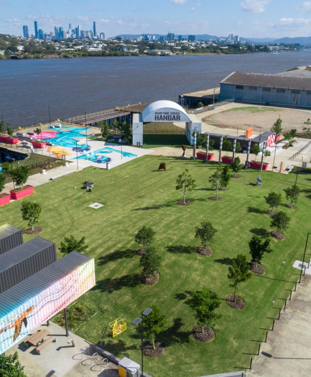 Aerial view of a riverside park with green lawns and scattered trees, showing the Maritime Green Hangar structure.