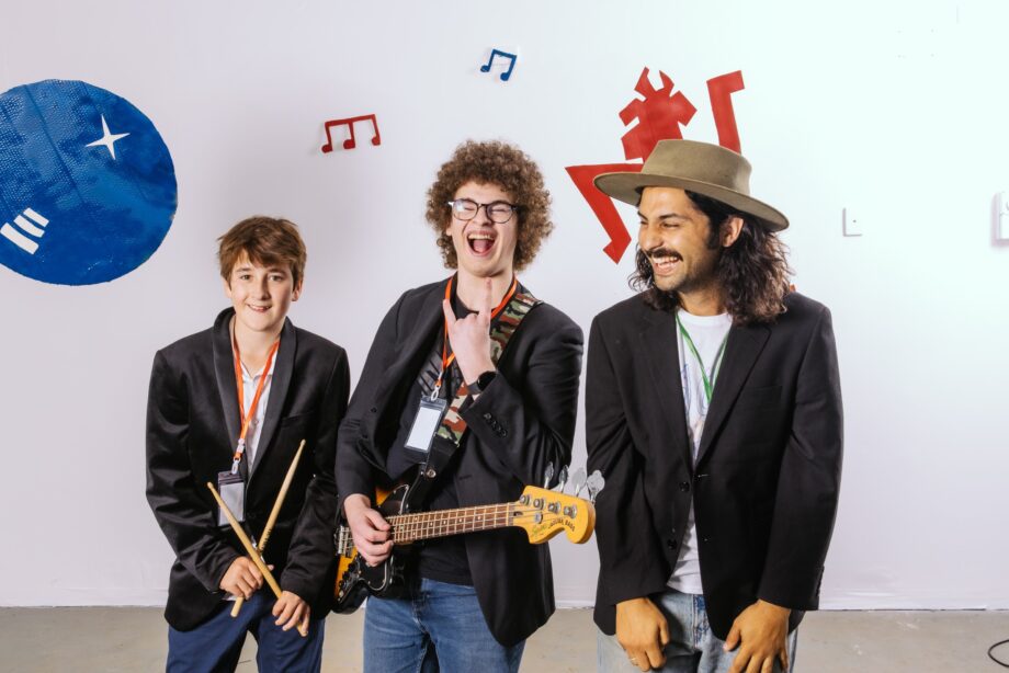 Three people in black jackets smile and play musical instruments in a fun, colorful room. One holds drumsticks, another a bass guitar, and the third stands with hands in pockets. Music notes and vibrant shapes decorate the white walls behind them.