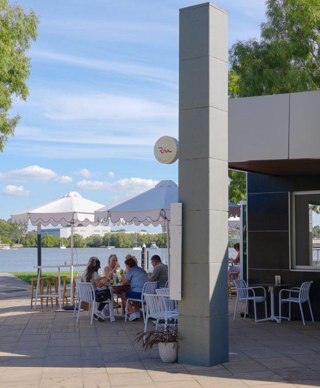 Outdoor dining at Riva, with several people sitting at tables under umbrellas near the Brisbane river.