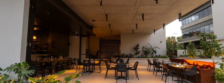 Outdoor dining at Ach Wine Bar with modern black chairs and tables, lit by ceiling lights. Surrounded by greenery and plants, the setting has a contemporary and inviting atmosphere.