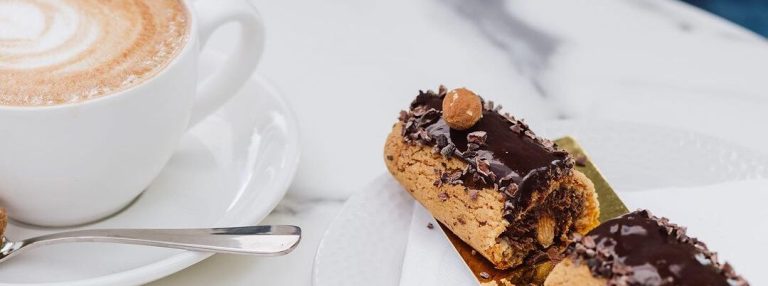 A cup of cappuccino sits on a marble table beside a white plate with a sliced chocolate pastry at Rise Bakery.