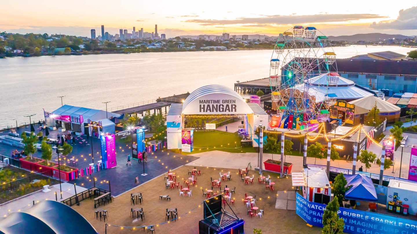 A vibrant riverfront scene at sunset featuring a Ferris wheel, colourful tents, and a central building labeled Maritime Green Hangar.