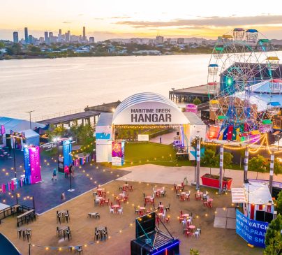 A vibrant riverfront scene at sunset featuring a Ferris wheel, colourful tents, and a central building labeled Maritime Green Hangar.