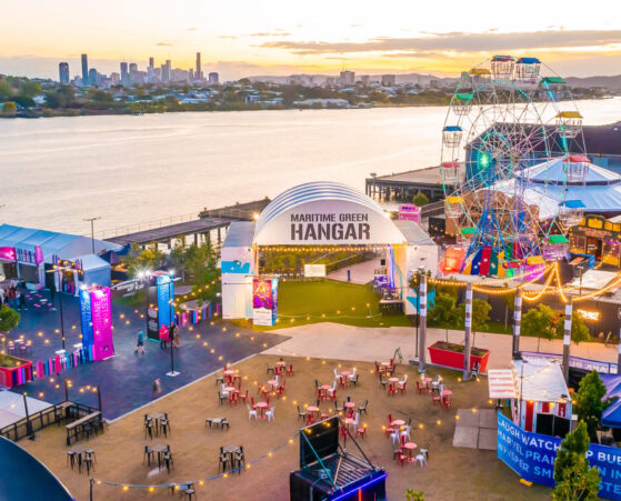 A vibrant riverfront scene at sunset featuring a Ferris wheel, colourful tents, and a central building labeled Maritime Green Hangar.