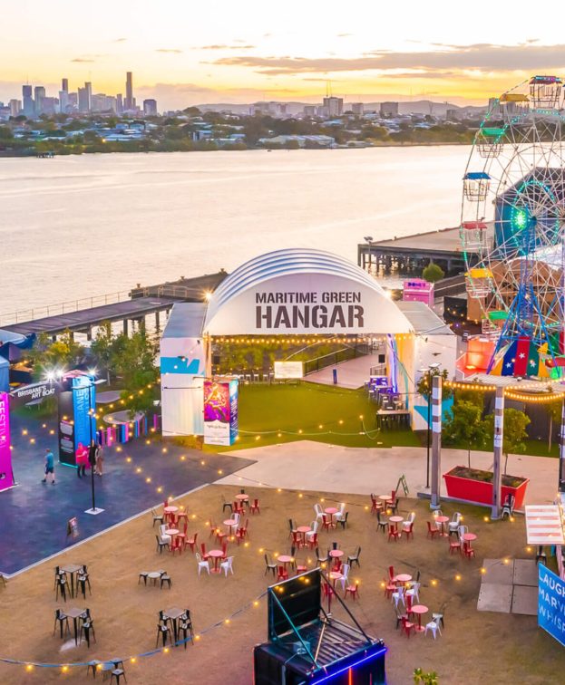 A vibrant riverfront scene at sunset featuring a Ferris wheel, colourful tents, and a central building labeled Maritime Green Hangar.
