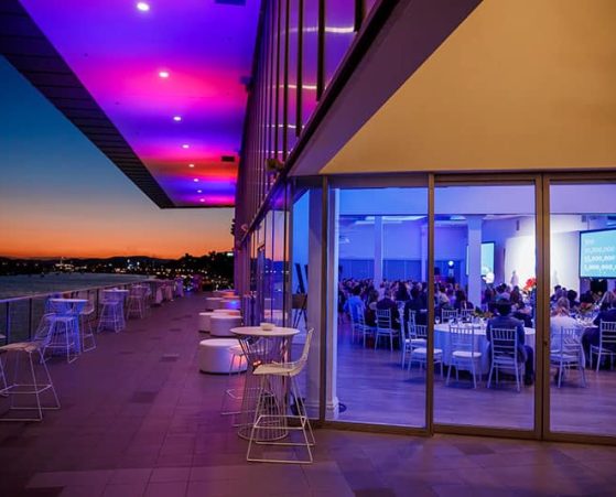 An elegantly lit venues with large windows reveals guests seated at tables during an event.