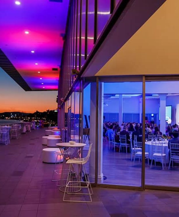 An elegantly lit venues with large windows reveals guests seated at tables during an event.