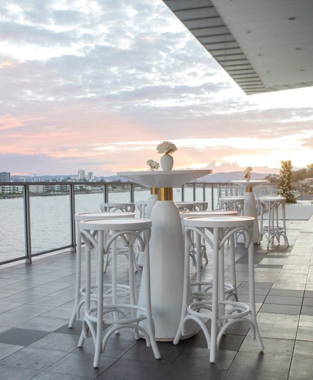 An outdoor seating area at Waters Edge with white bar tables and stools overlooks a waterfront cityscape at sunset.