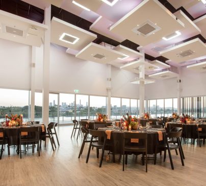 A spacious, elegantly decorated banquet hall with large windows showcasing a cityscape. Smooth wooden floors, round tables with black chairs, and floral centerpieces create a sophisticated atmosphere. Ceiling lights in modern square designs add to the ambiance.