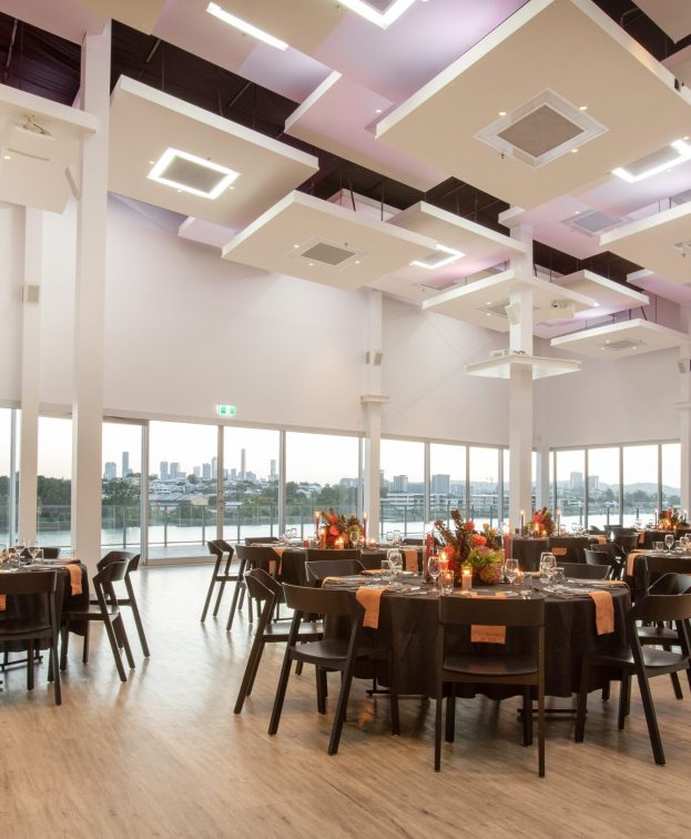 A spacious, elegantly decorated banquet hall with large windows showcasing a cityscape. Smooth wooden floors, round tables with black chairs, and floral centerpieces create a sophisticated atmosphere. Ceiling lights in modern square designs add to the ambiance.