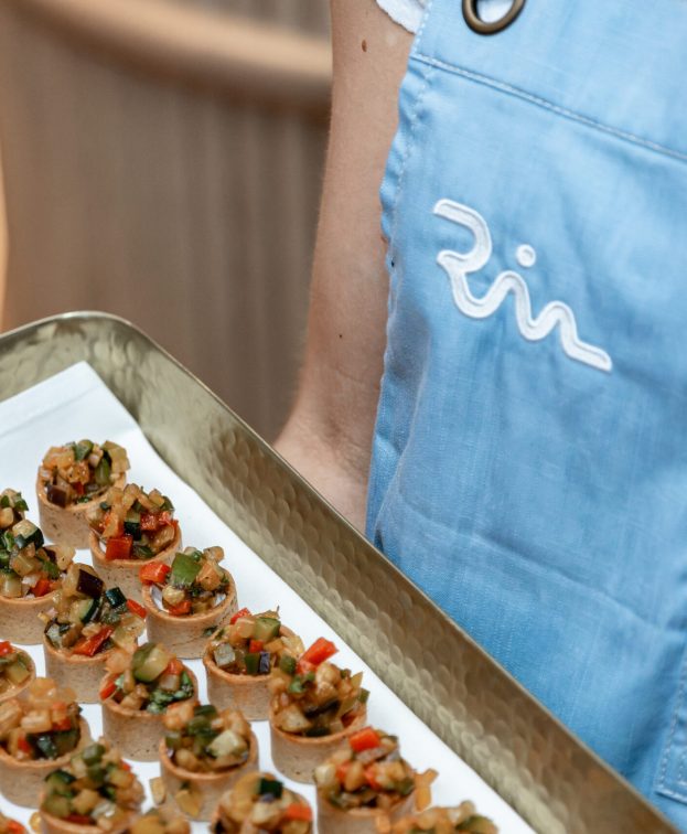 A person in a light blue Riva apron holds a tray of appetizers.