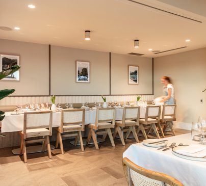 A well-lit dining room at Riva, with a long table set for a meal, adorned with white tablecloths, plates, and cutlery.