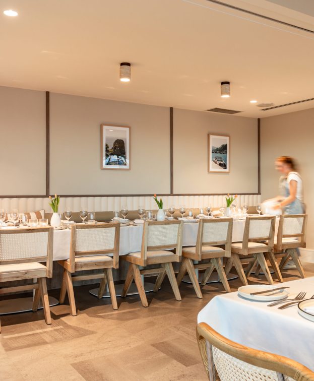 A well-lit dining room at Riva, with a long table set for a meal, adorned with white tablecloths, plates, and cutlery.