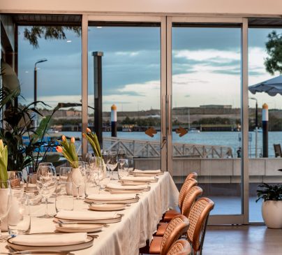 Elegant dining setup with a long table covered in a white tablecloth, set with glassware and tulips in vases. Large glass doors open to a scenic waterfront view with trees and a patio, under a partly cloudy sky.