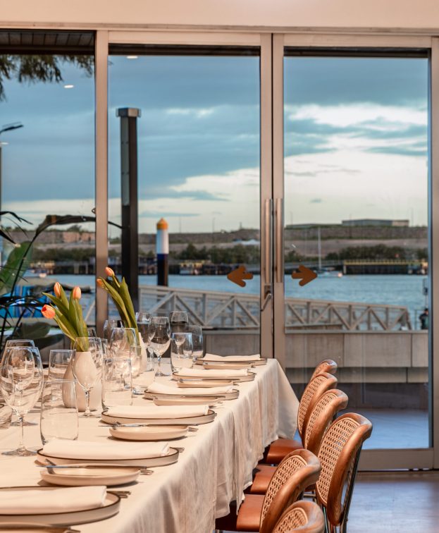 Elegant dining setup with a long table covered in a white tablecloth, set with glassware and tulips in vases. Large glass doors open to a scenic waterfront view with trees and a patio, under a partly cloudy sky.
