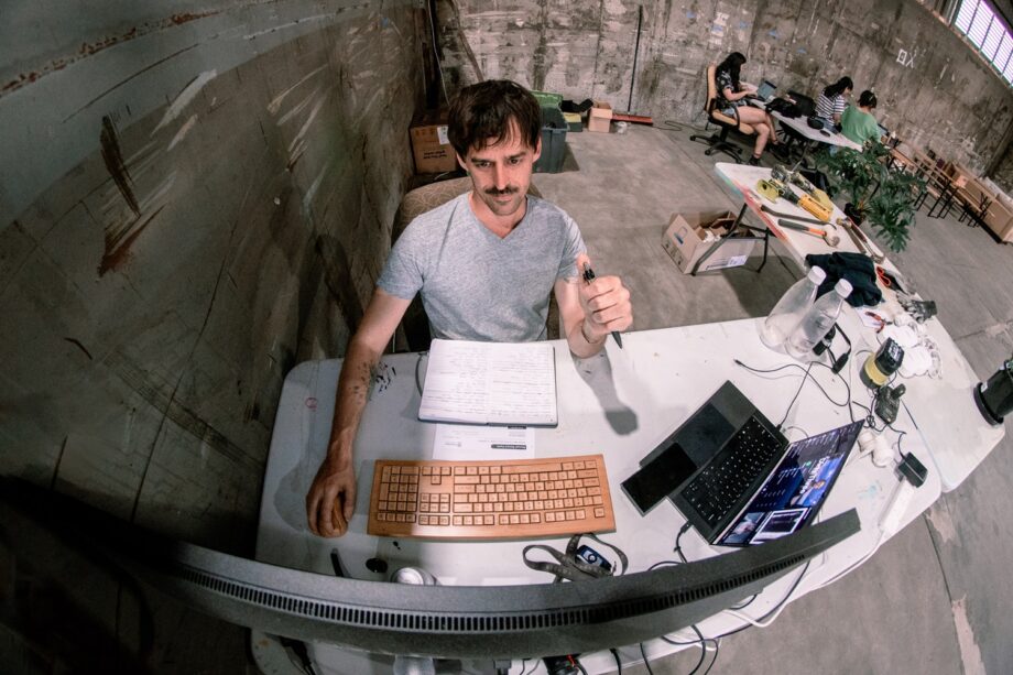 A man with a mustache sits at a cluttered desk in an industrial-style room, working on a computer and taking notes. Other people are visible in the background, and the space has a concrete wall and an open, creative atmosphere.