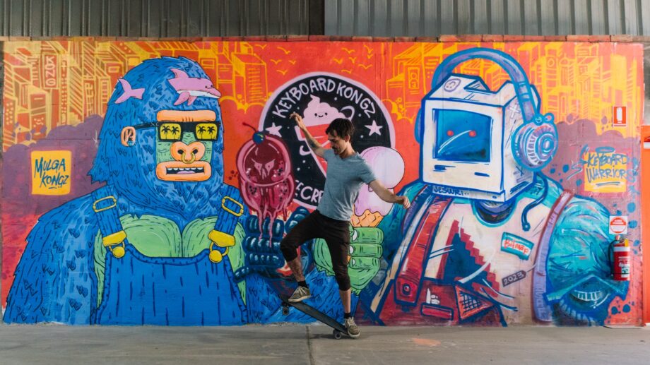 A person skateboards in front of a colorful graffiti wall featuring a gorilla with sunglasses, a retro computer, and vibrant, abstract designs.