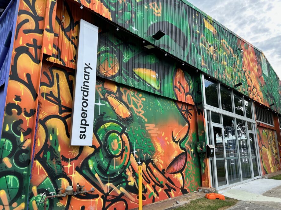 A vibrant street art mural covers the side of a building, featuring abstract designs in green, orange, and yellow. A white sign with black text reads superordinary. Large glass doors provide an entrance.
