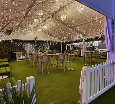 A festive outdoor marquee decorated with strings of warm white fairy lights. Several bar stools and high tables are arranged under the marquee, surrounded by white picket fencing and greenery.