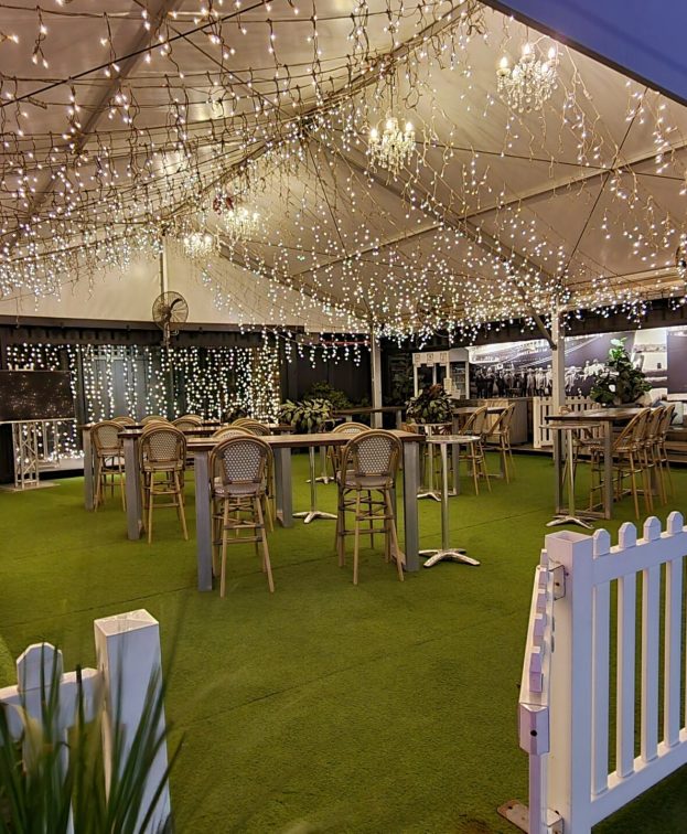 A festive outdoor marquee decorated with strings of warm white fairy lights. Several bar stools and high tables are arranged under the marquee, surrounded by white picket fencing and greenery.