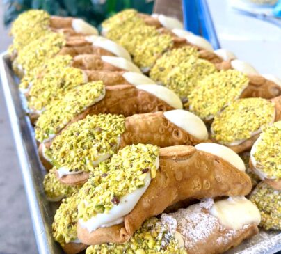 A tray filled with multiple cannoli, each topped with chopped pistachios and filled with creamy filling.