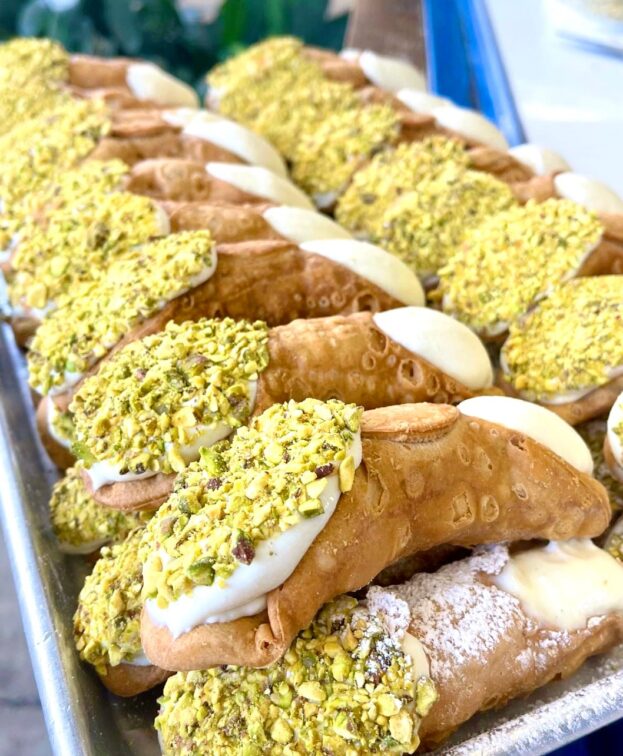 A tray filled with multiple cannoli, each topped with chopped pistachios and filled with creamy filling.
