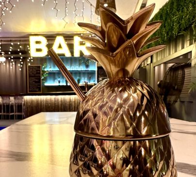 A shiny gold pineapple-shaped cocktail cup with a straw sits on a marble counter. In the background, a bar sign is illuminated with string lights, creating a cozy and inviting atmosphere.