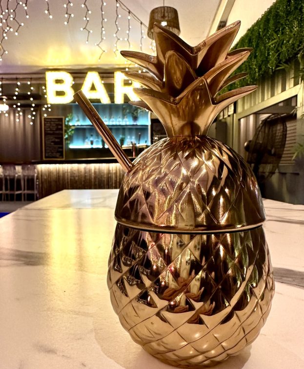 A shiny gold pineapple-shaped cocktail cup with a straw sits on a marble counter. In the background, a bar sign is illuminated with string lights, creating a cozy and inviting atmosphere.