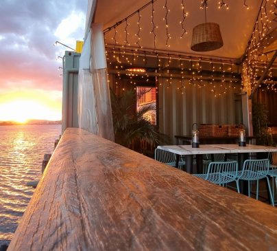 The Eat Street River Deck overlooking the river at sunset. The warm glow of string lights and a BAR sign create a cozy atmosphere. The horizon features a vibrant sky with clouds reflecting the sunsets colors.