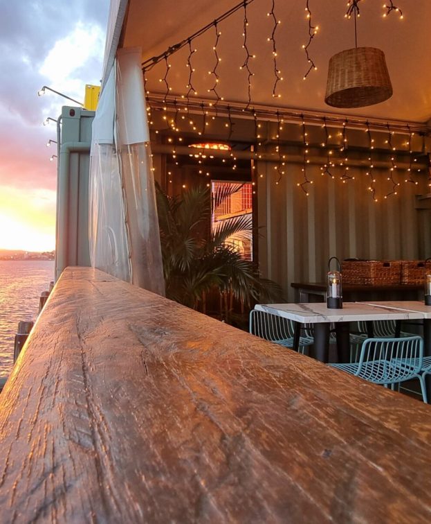 The Eat Street River Deck overlooking the river at sunset. The warm glow of string lights and a BAR sign create a cozy atmosphere. The horizon features a vibrant sky with clouds reflecting the sunsets colors.