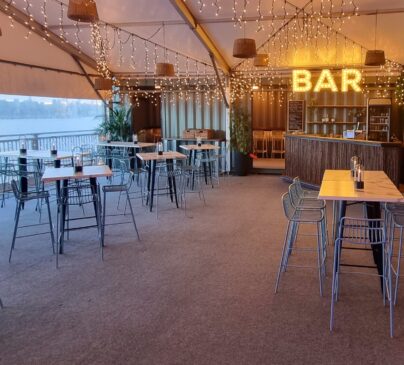 Eat Street River Deck's function space. High tables and stools are arranged across the floor. A bar sign and fairy lights glow in the background, with a view of water outside.