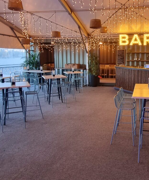 Eat Street River Deck's function space. High tables and stools are arranged across the floor. A bar sign and fairy lights glow in the background, with a view of water outside.