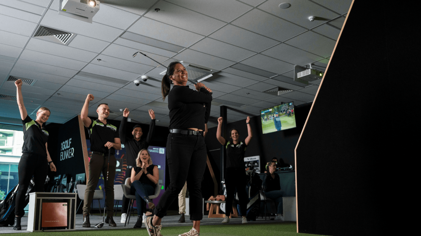 A group of people celebrates indoors as a woman in the foreground poses with a golf club, simulating a swing. They are on a golf simulator platform, and the room has high ceilings and equipment around.