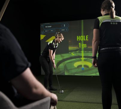 One person is about to putt on an artificial green, while others watch. A digital screen displays the phrase HOLE 1 m and other golf game details.