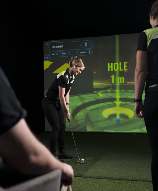 One person is about to putt on an artificial green, while others watch. A digital screen displays the phrase HOLE 1 m and other golf game details.
