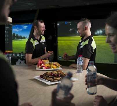 Two people enjoy catered fruit and snacks in front of the golf simulator.