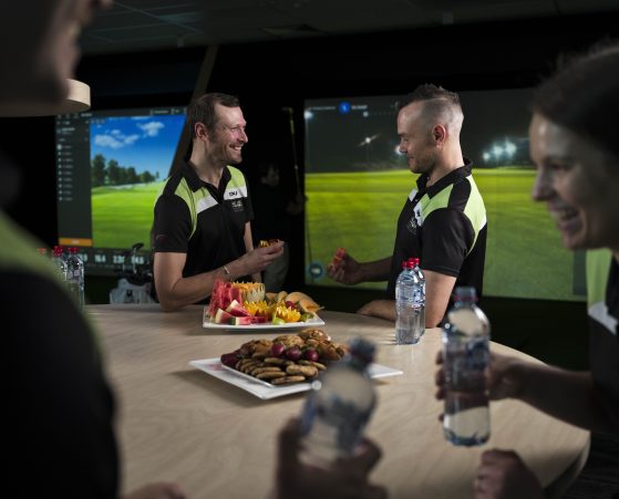 Two people enjoy catered fruit and snacks in front of the golf simulator.
