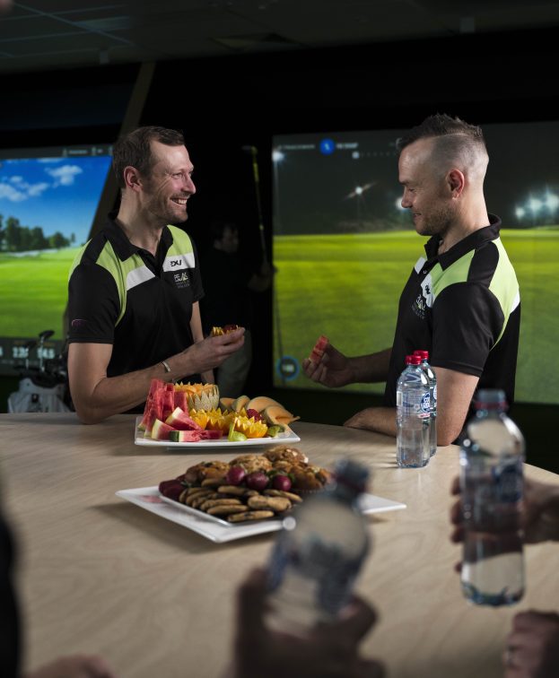 Two people enjoy catered fruit and snacks in front of the golf simulator.