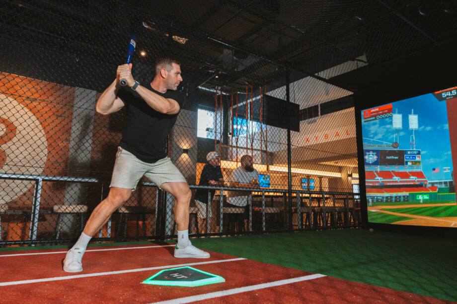A person in casual clothing is standing in a batting stance inside a virtual batting cage, holding a baseball bat. A digital screen shows a baseball field, and people are visible in the background observing.