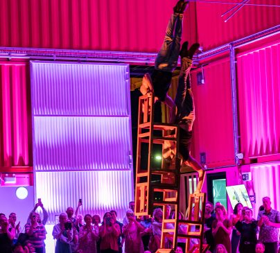 An acrobat performs an impressive balance act on a stack of chairs, elevated and upside down, in a brightly lit, colourful venue. A crowd watches in awe