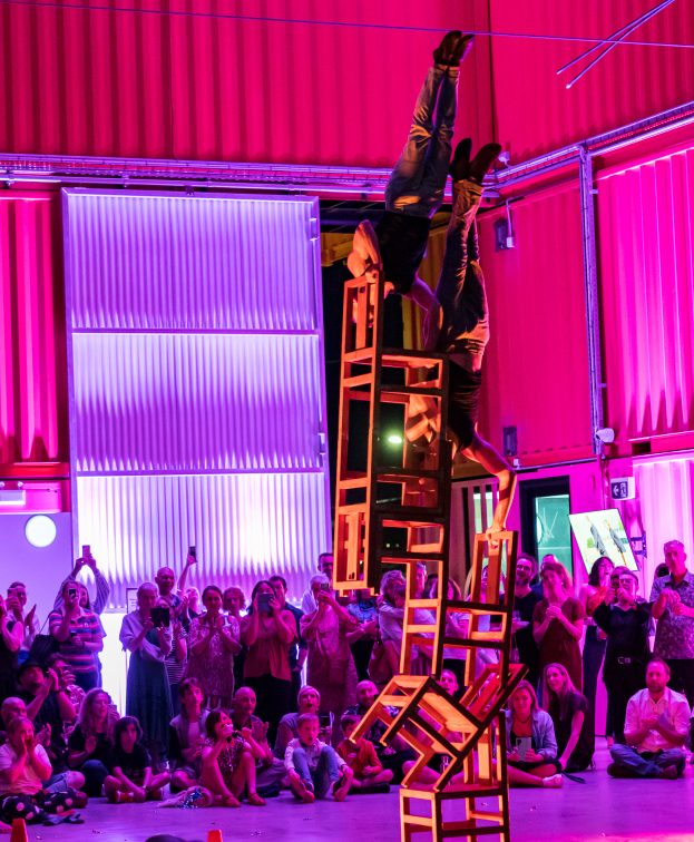 An acrobat performs an impressive balance act on a stack of chairs, elevated and upside down, in a brightly lit, colourful venue. A crowd watches in awe